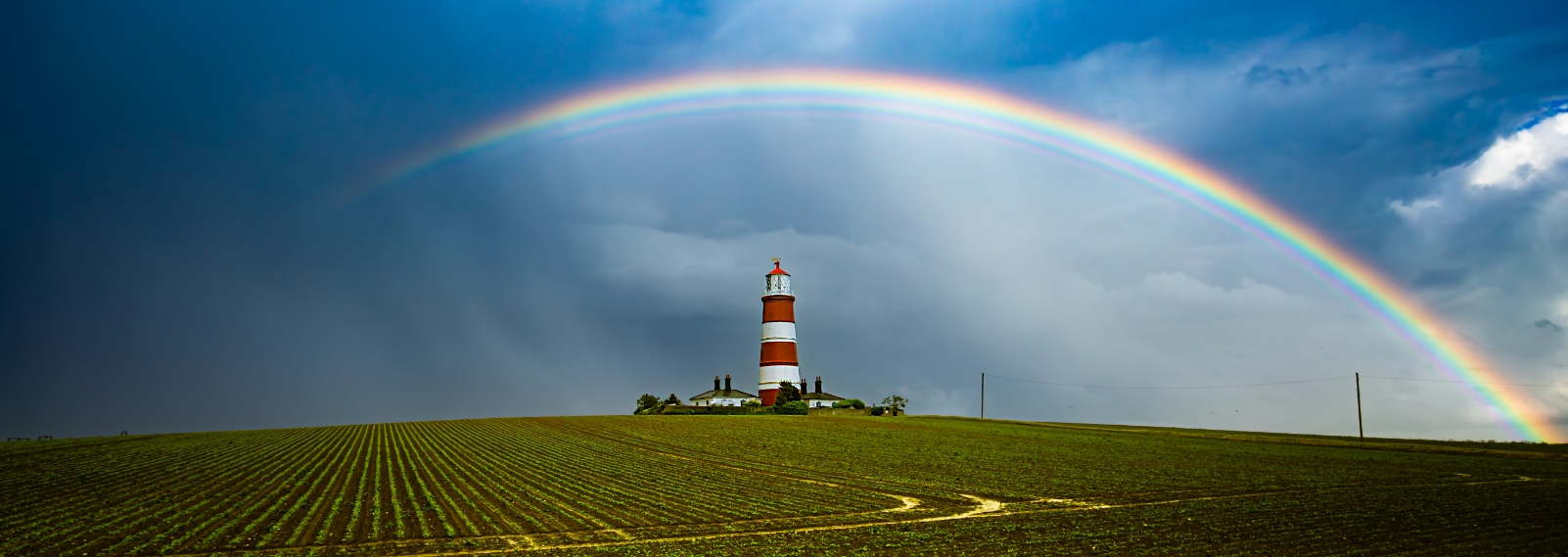 Ein Leuchtturm am Horizont mit Regenbogen als Titelbild für 'Über uns'
